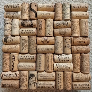 May include: A square arrangement of numerous light brown natural corks, each featuring various black printed brand names, logos, and text such as In Barrel We Toast, King Stag, Clay Creek, and 19 Crimes, creating a textured grid pattern.