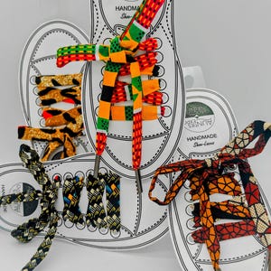 May include: Handmade shoe laces in various colorful African-inspired patterns. The laces are displayed on white shoe-shaped cards with the text "Aunties Expo Handmade Shoe-Laces". The laces feature vibrant colors like red, orange, yellow, and black.