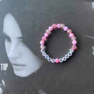 May include: A pink and white beaded bracelet with the words "VIDEO GAMES" spelled out in white beads.