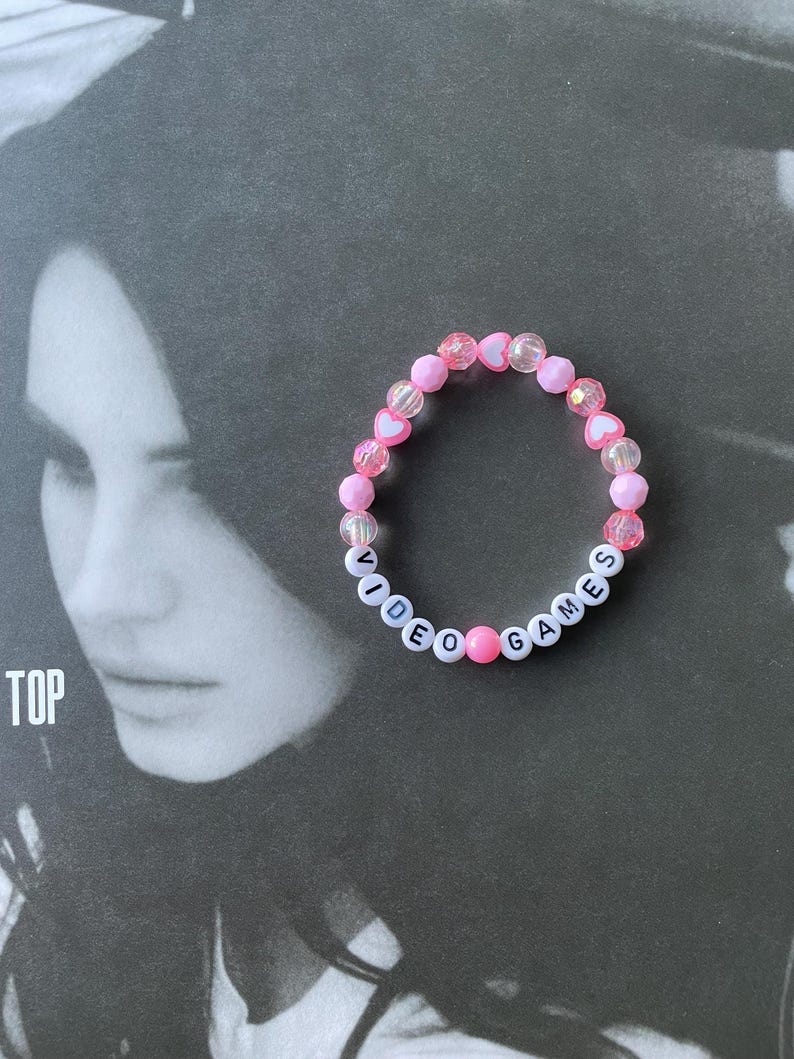 May include: A pink and white beaded bracelet with the words "VIDEO GAMES" spelled out in white beads.