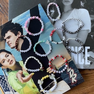 May include: A collection of colorful beaded bracelets with different words spelled out on them, including "Say Yes To Heaven", "Ultraviolence", "Happiness Is", "Cherry", "Star Girl", "Pretty When You Cry", "Lana Del Rey", "Cinnamon Girl", and "Let The Light In".