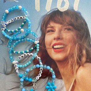 May include: A stack of blue beaded bracelets with white lettering spelling out "Say You The Game", "Still Over", "Blue Dress", "No One", "Boat", and "1989".