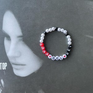 May include: A white beaded bracelet with black and red beads spelling out "Lana Del Rey" with red heart beads between the words.