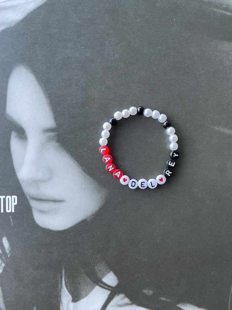 May include: A white beaded bracelet with black and red beads spelling out "Lana Del Rey" with red heart beads between the words.
