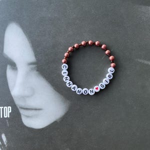May include: A beaded bracelet with the words "Cinnamon Girl" and a small red heart charm. The beads are a rose gold color.