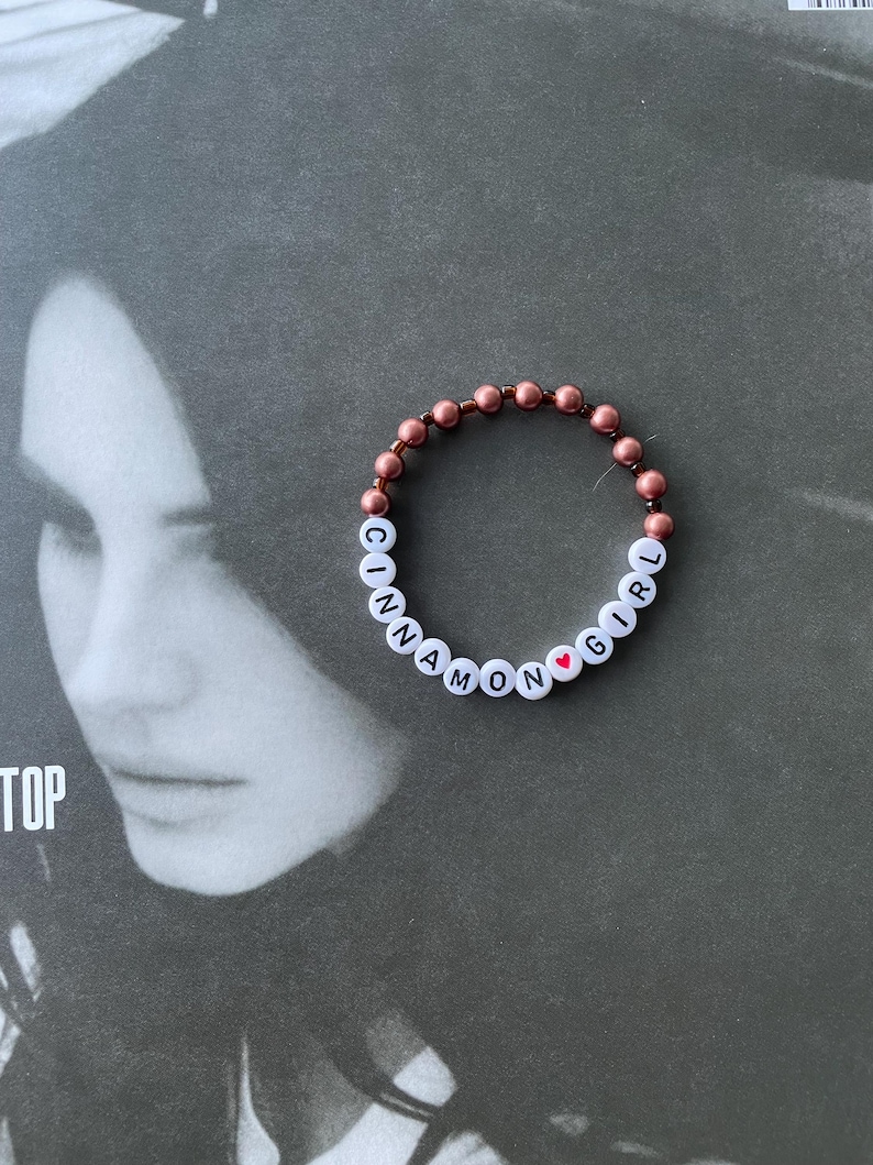 May include: A beaded bracelet with the words "Cinnamon Girl" and a small red heart charm. The beads are a rose gold color.
