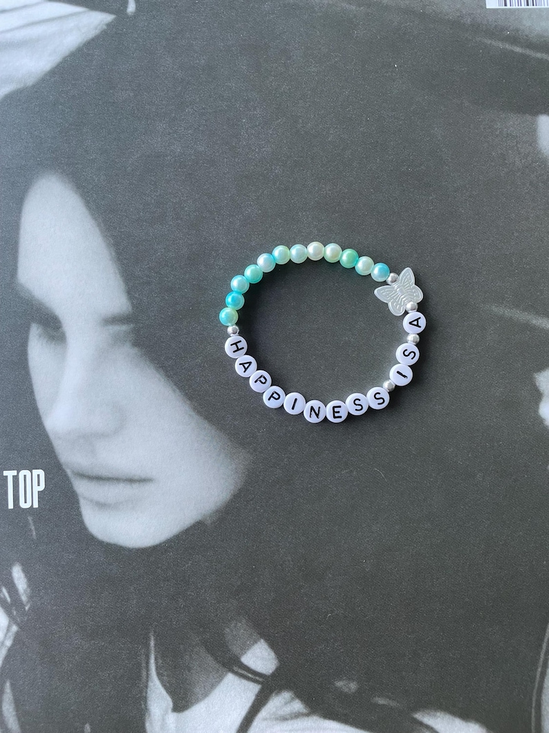 May include: A light blue and white beaded bracelet with the words "Happiness is a" spelled out in black letters. A silver butterfly charm is attached to the bracelet.