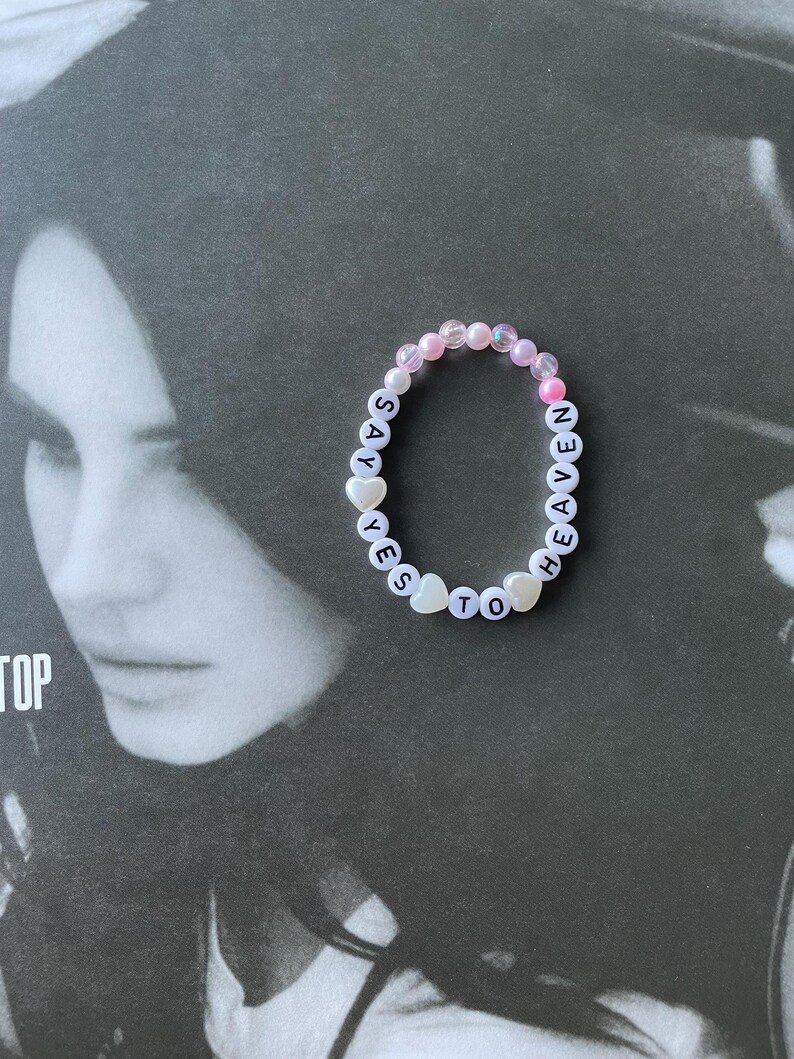 May include: A white beaded bracelet with the words "SAY YES TO HEAVEN" spelled out in black letters. The beads are pink, white, and iridescent.