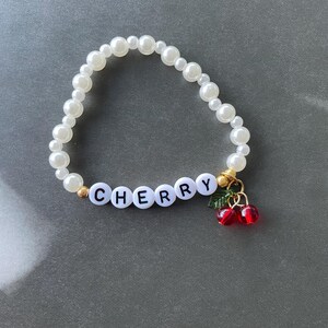 May include: A white pearl bracelet with the word "CHERRY" spelled out in black letters and a charm of two red cherries with green leaves.