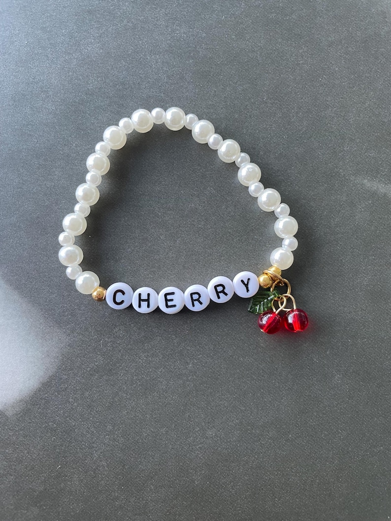 May include: A white pearl bracelet with the word "CHERRY" spelled out in black letters and a charm of two red cherries with green leaves.