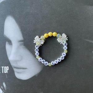 May include: A white beaded bracelet with the words "Let the Light In" spelled out in black letters. The beads are interspersed with yellow and clear plastic beads.