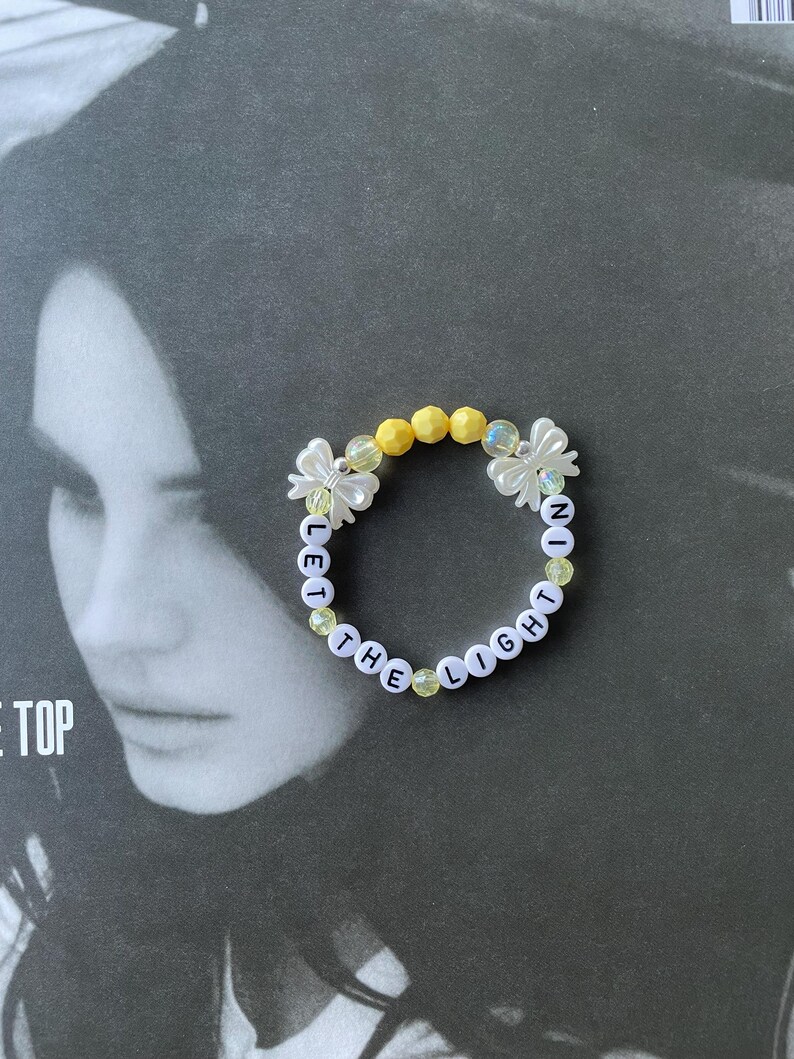 May include: A white beaded bracelet with the words "Let the Light In" spelled out in black letters. The beads are interspersed with yellow and clear plastic beads.