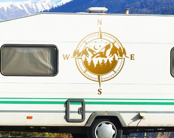 May include: A white camper van with a gold compass decal featuring a mountain range and pine trees. The compass points north, south, east, and west.
