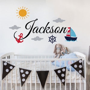 May include: A white cot with a black and white polka dot bunting hanging above it. The wall behind the cot has a nautical themed decal with a sailboat, anchor, steering wheel, clouds, and the name "Jackson".