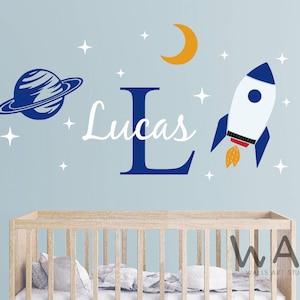 Könnte beinhalten: Ein Wandaufkleber-Set mit einer blauen und weißen Rakete, einem blauen Planeten mit Ring, einem gelben Halbmond und weißen Sternen. Der Name "Lucas" ist in weißen Kursivbuchstaben mit einem blauen Buchstaben "L" geschrieben.