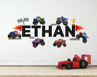 May include: A black and white wall decal with the name "ETHAN" spelled out in large letters. The letters are decorated with a black road design. There are several monster truck decals around the name, including a purple monster truck with flames, a green monster truck with a checkered flag, a blue monster truck with a trophy, and a red monster truck with a checkered flag.