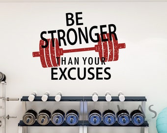 May include: A white wall with a red and black motivational quote that reads "Be Stronger Than Your Excuses" with a red barbell graphic. The quote is on a white wall with a rack of dumbbells in the foreground.