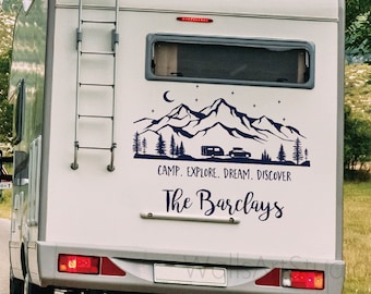 Puede incluir: Una autocaravana blanca con una calcomanía de una escena de montaña azul en la parte trasera. La calcomanía incluye una luna creciente, estrellas, árboles y una pequeña autocaravana. El texto "Camp. Explore. Dream. Discover" está debajo de la escena, y el texto "The Barclays" está debajo de eso.
