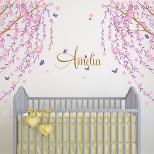 May include: A nursery wall decal featuring two weeping willow trees with pink flowers and purple butterflies. The name "Amelia" is written in cursive script on the wall.