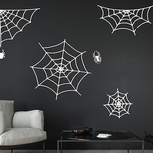 May include: Five white spiderweb wall decals on a black wall. The spiderwebs are various sizes and shapes. Two small white spiders are also included.
