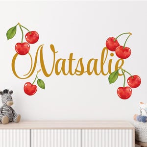 Puede incluir: Una calcomanía de pared de vinilo dorado con el nombre "Watsalie" escrito en letra cursiva. El nombre está rodeado de cerezas rojas con hojas verdes.