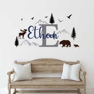 May include: A grey letter E with the name Ethan written in blue script above it. The letter E is surrounded by mountains, trees, a moose, and two bears. The design is on a white wall above a wooden bench with two pillows.