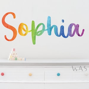 Puede incluir: Un vinilo decorativo de pared con el nombre "Sophia" en cursiva. Las letras cambian de rojo a morado. El vinilo está sobre una cómoda blanca con pomos de colores y bloques de madera.
