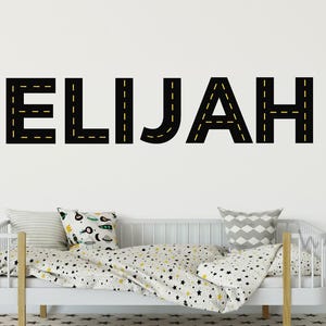 Op de afbeelding: Zwarte en gele wanddecoratie met een wegthema die de naam "ELIJAH" spelt. De sticker zit op een witte muur boven een wit dagbed met een wit en zwart beddengoed met sterrenpatroon.