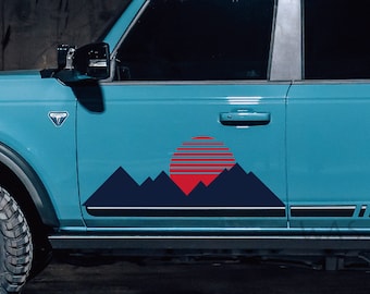 May include: A blue Ford Bronco with a black and white mountain and sunset graphic decal on the side door. The decal features a red sun with stripes and blue mountains.
