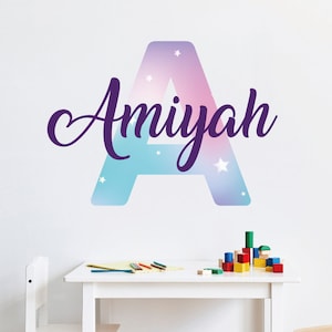Puede incluir: Una letra A azul y rosa pastel con estrellas blancas, con el nombre "Amiyah" escrito en cursiva morada.