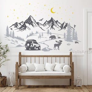 Op de afbeelding: Een muursticker met een berglandschap met een voertuig, dieren in het wild en hemellichamen. Het ontwerp omvat een bergketen, bomen, een berenfamilie, een eland en een halve maan. Het kleurenschema is voornamelijk grijs, zwart en geel.
