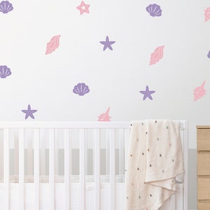 Könnte beinhalten: Ein weißes Babybett mit einer weißen Decke und einer weißen Wand mit violetten und rosa Muscheln und Seestern-Aufklebern.