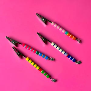 May include: Four metal alligator clips with colorful, striped handles. The handles feature a variety of colors, including pink, blue, green, orange, and yellow, against a bright pink background. Each clip has a small, heart-shaped detail.