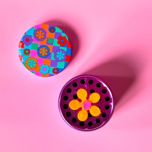 May include: Purple metal grinder with a flower design on the lid. The lid has a checkered pattern with colorful flowers. The grinder has a flower-shaped screen with a pink center.