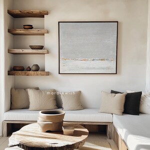 May include: A light gray abstract painting with gold accents hangs above a white couch with beige and gray pillows. The couch is in a room with wooden shelves and a wooden coffee table.