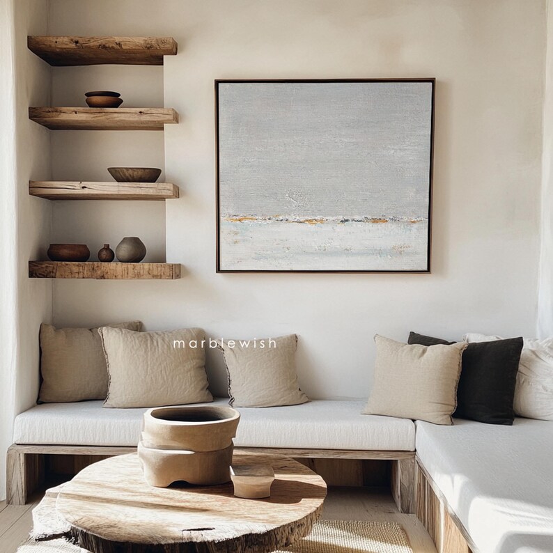May include: A light gray abstract painting with gold accents hangs above a white couch with beige and gray pillows. The couch is in a room with wooden shelves and a wooden coffee table.