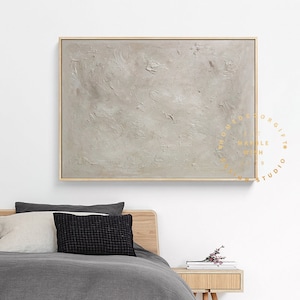 May include: A large, rectangular abstract painting with a textured, light gray surface. The artwork is framed in a thin, natural wood frame. The painting is displayed above a bed with gray bedding and pillows.