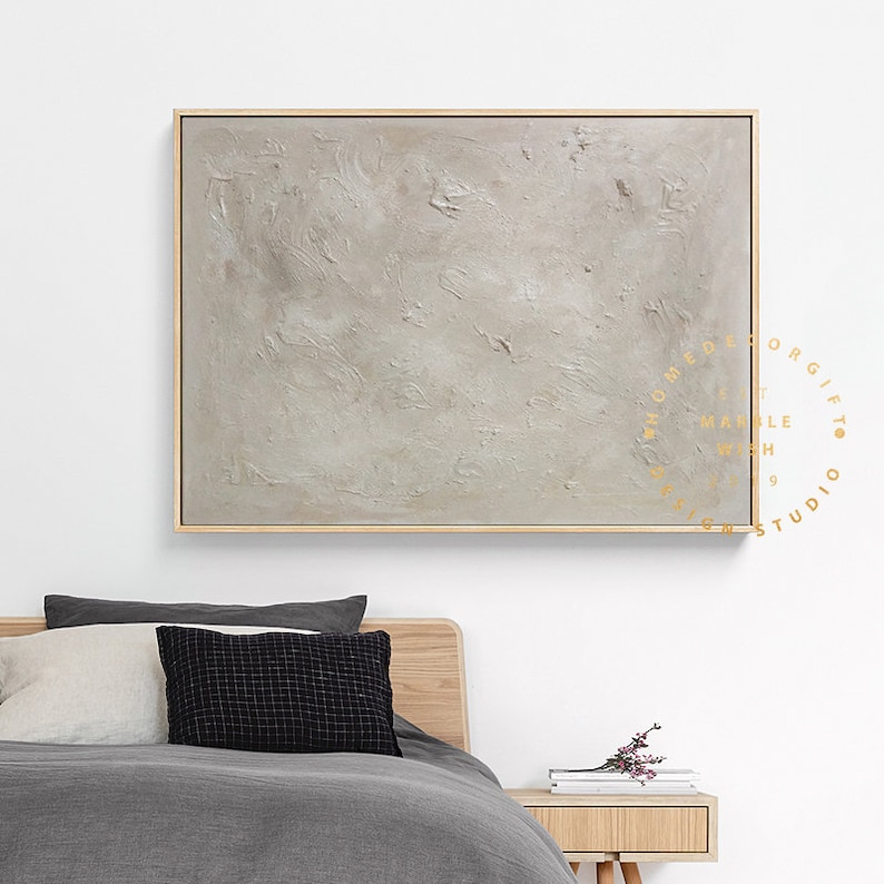 May include: A large, rectangular abstract painting with a textured, light gray surface. The artwork is framed in a thin, natural wood frame. The painting is displayed above a bed with gray bedding and pillows.