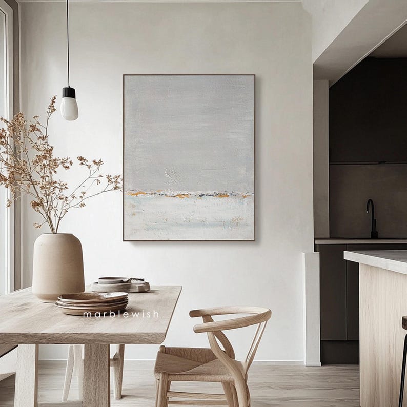 May include: A minimalist abstract painting with a gray and white background and a thin line of gold paint across the middle. The painting is hanging on a white wall in a dining room with a wooden table and chairs.
