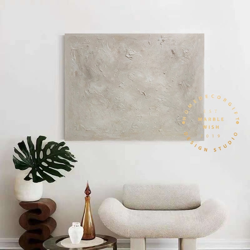 May include: A large, textured, neutral-toned abstract painting hangs above a modern, curved white chair. A dark wooden side table holds a white vase with a large green leaf. A small, round, metal side table holds a brown glass bottle and a white vase.