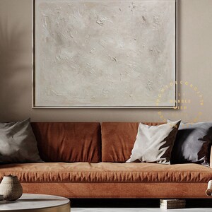 May include: A large, square abstract painting with a textured, off-white surface, framed in a thin gold border. The artwork hangs above a rust-colored sofa with gray pillows, creating a warm, inviting living room scene.