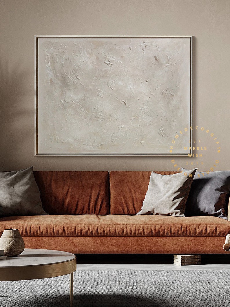 May include: A large, square abstract painting with a textured, off-white surface, framed in a thin gold border. The artwork hangs above a rust-colored sofa with gray pillows, creating a warm, inviting living room scene.