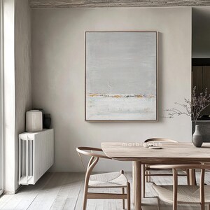 May include: Abstract painting in shades of gray and gold with a subtle horizontal line in the center. The painting is framed and hanging on a wall in a modern dining room setting.