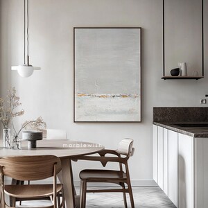 May include: A gray and white abstract painting with gold accents hangs on a white wall above a wooden dining table with chairs. The painting has a horizontal line of gold paint across the middle.