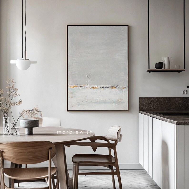 May include: A gray and white abstract painting with gold accents hangs on a white wall above a wooden dining table with chairs. The painting has a horizontal line of gold paint across the middle.