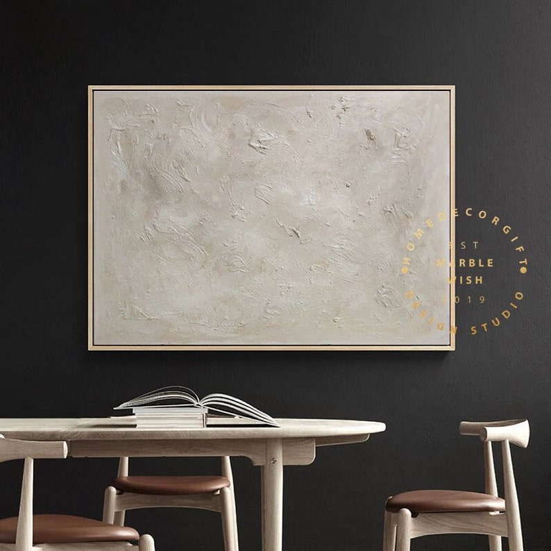 May include: Abstract beige textured wall art in a gold frame. The artwork has a neutral color palette and a minimalist design. The painting is displayed above a wooden table with chairs, creating a modern interior design.