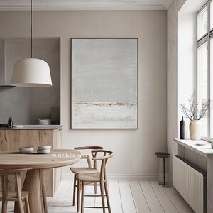 May include: Abstract landscape painting with a gray background, white and gold accents, and a subtle blue horizon line. The painting is framed and hanging on a wall in a modern dining room.