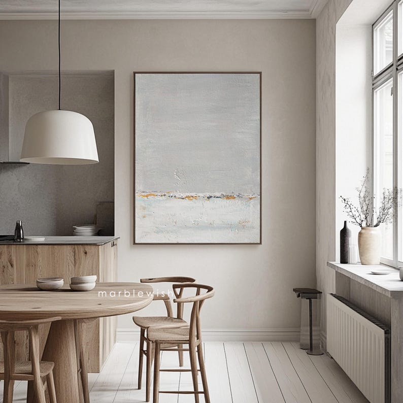 May include: Abstract landscape painting with a gray background, white and gold accents, and a subtle blue horizon line. The painting is framed and hanging on a wall in a modern dining room.