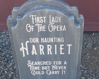 May include: A grey tombstone with white lettering that reads "First Lady of the Opera, Our Haunting Harriet, Searched for a Tune but Never Could Carry It."
