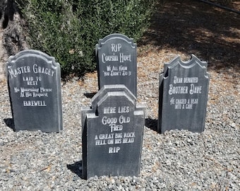 May include: Four gray tombstone-shaped decorations with humorous epitaphs. The tombstones are on a bed of gray gravel.